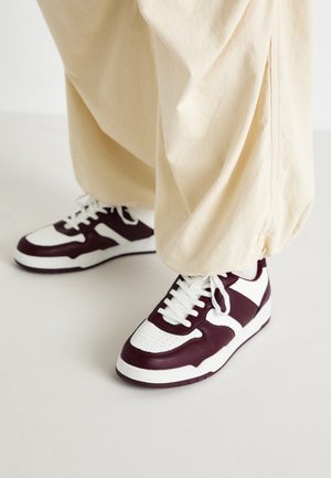 Person iført cremegule, vide bukser og hvide sneakers med mørkebrune detaljer og hvide snørebånd, stående på et lyst gulv.