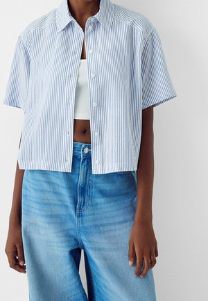 Personne portant une chemise à manches courtes rayée bleue et blanche boutonnée sur un crop top blanc et un jean taille haute bleu clair.