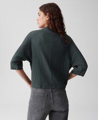 Vrouw die wegkijkt, gekleed in een gestructureerde donkergroene top met driekwartmouwen en vervaagde zwarte jeans, met de handen op de heupen.