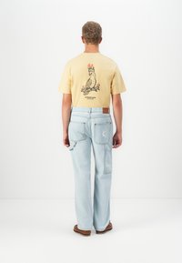 Muž s krátkymi kučeravými vlasmi, ktorý má oblečené žlté tričko s grafickým motívom plameňového vtáka a svetlomodré cargo jeans, stojí proti bielej stene.