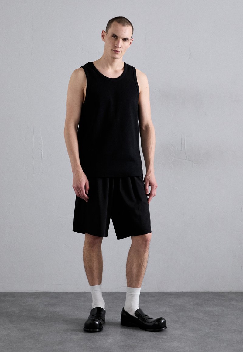 Schwarzes ärmelloses Tanktop und lockere schwarze Shorts aus Stoff, kombiniert mit schwarzen Schuhen und weißen Socken, stehend vor einem grauen Hintergrund.