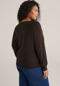 Hnědý pletený svetr s volným střihem, dlouhými rukávy a ribbovaným lemem. Nosený s modrými džíny, zobrazující zadní pohled na design.