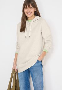 Femme souriante portant un sweat à capuche beige et un jean bleu, tenant une veste kaki dans une main, l'autre main dans sa poche, debout contre un mur blanc.