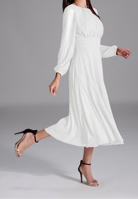 Robe blanche à manches longues avec un corsage ajusté et une jupe fluide. Sandales à talons hauts noires avec des lani ères et des accents dorés. Texture de tissu lisse.