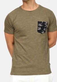 Camiseta de color verde oliva con textura moteada, cuello redondo y un bolsillo negro con un detalle de impresión de un pájaro blanco. Manga corta.