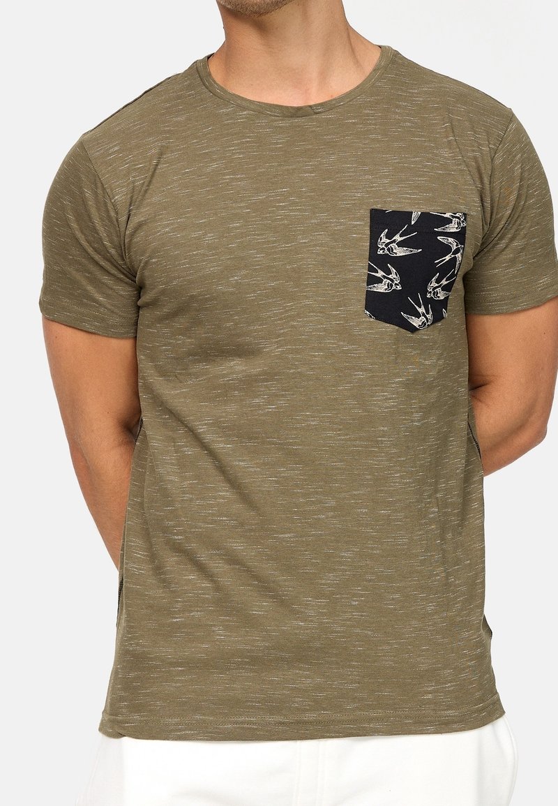 Olivgrünes T-Shirt mit einer gesprenkelten Textur, rundem Halsausschnitt und einer schwarzen Tasche mit einem weißen Vogel-Print-Detail. Kurze Ärmel.