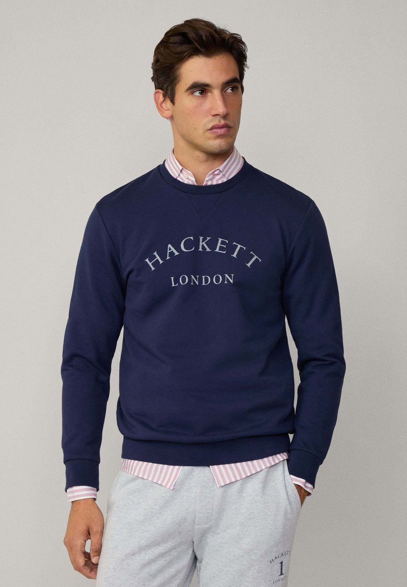 Marineblå sweatshirt med "HACKETT LONDON" i lysegrå skrift, rund hals og ribbede cuffe. Båret over en lyserød stribet skjorte.