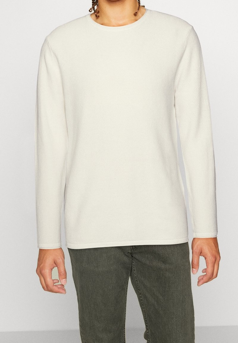 Kremowa koszulka z długim rękawem wykonana z teksturowanej tkaniny, z zaokrąglonym dekoltem i kontrastowym obszyciem na mankietach i dole.