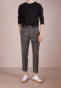Schwarzes Langarmshirt, graue Plissee-Hose, schwarzer Leder-Gürtel und weiße Sneakers mit braunen Sohlen, die ein lässiges Outfit präsentieren.