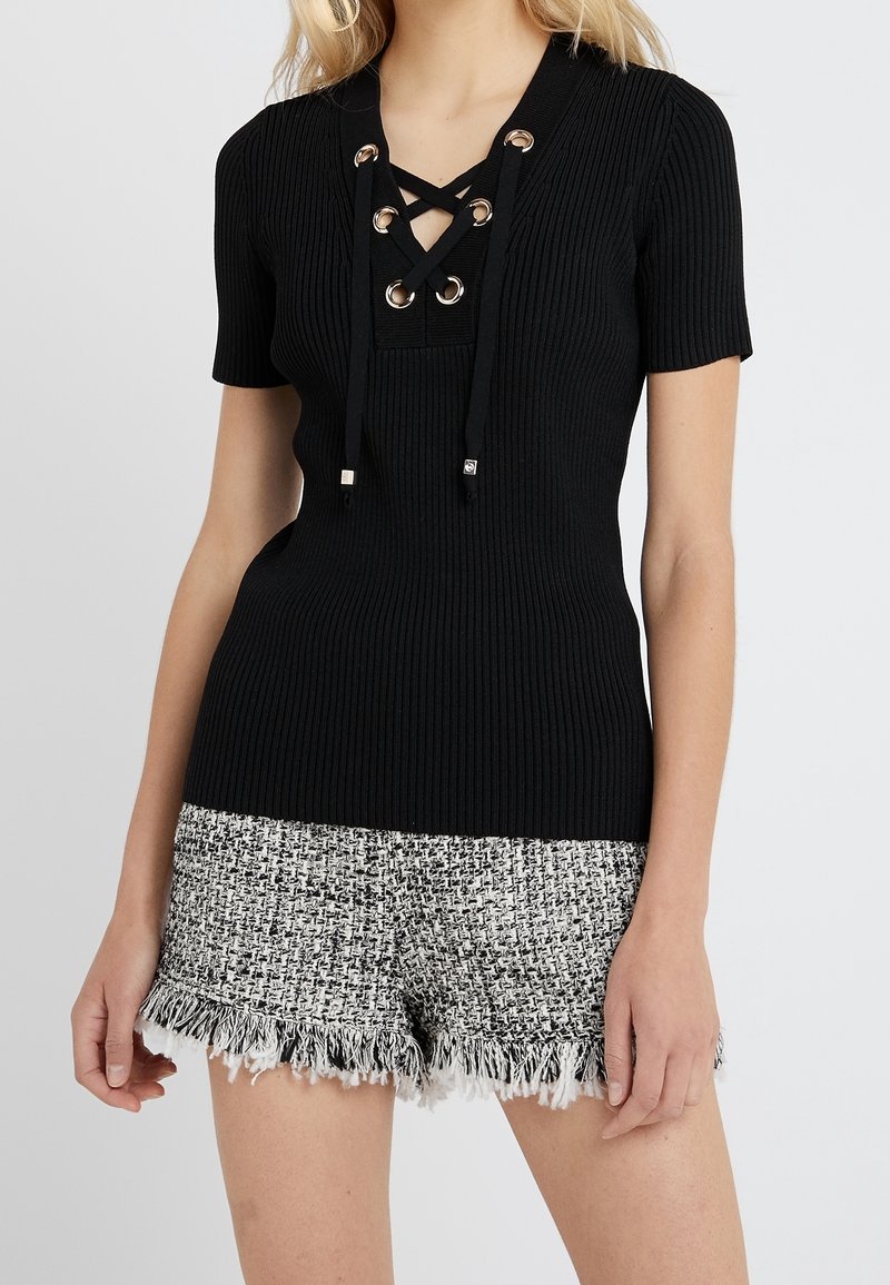 Femme portant un haut noir côtelé à manches courtes avec un col lacé en croisillon et un short en tweed noir et blanc à ourlet effiloché.