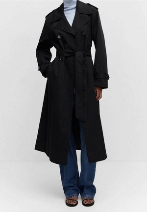 Manteau trench noir long avec ceinture nouée à la taille, porté sur un col roulé bleu clair, associé à un jean bleu et des sandales ouvertes.