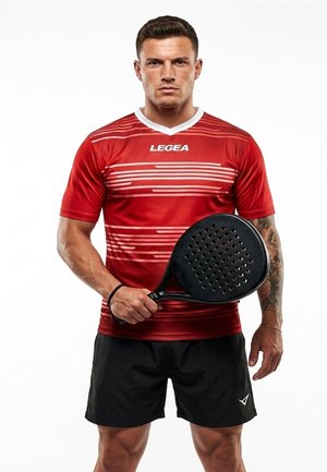 Uomo con maglietta sportiva rossa e pantaloncini neri che tiene una racchetta da padel nera, in piedi su sfondo bianco.