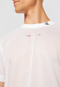 Bílý síťovaný tričko s kulatým výstřihem a krátkými rukávy. Na něm je vytištěn text "Tohle je láska" v duhových barvách a černé logo "CK" na límci.