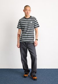 Pruhované černo-šedé tričko s malým bílým logem, doplněné tmavě šedými džíny a teniskami s oranžovými akcenty na modrém koberci.