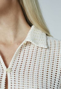 Hvid strikket bluse med krave, der har et rutemønster og knaplukning. Let og semi-transparent tekstur.