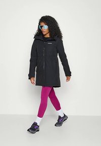 Woman with curly hair wearing oversized sunglasses, a black zip-up coat, purple leggings, white socks, and black sneakers walking.
