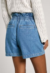Pantalones cortos de mezclilla con cintura elástica, color azul, corte holgado, dos bolsillos traseros y dobladillo limpio. Combinados con una blusa de punto blanca.