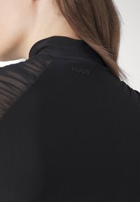 Gros plan sur une personne portant un haut noir à col montant avec des détails en mesh sur l'épaule, avec le logo "HUGO" au dos.