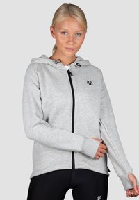 Svetlošedá mikina na zips s kapucňou, dlhými rukávmi a rebrovanými manžetami. Na hrudi má čierne logo a hladký povrch.