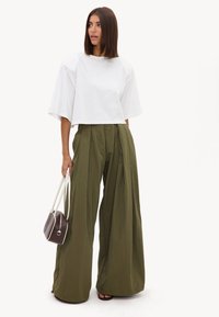 Femme aux cheveux bruns raides portant un haut blanc à manches courtes oversize et un pantalon plissé large kaki, tenant un petit sac à main marron.