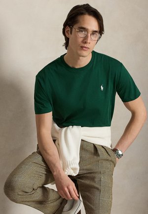 Ung mand med briller, grøn T-shirt, ternede bukser, hvid sweater bundet om livet, siddende med det ene ben løftet mod en neutral baggrund.