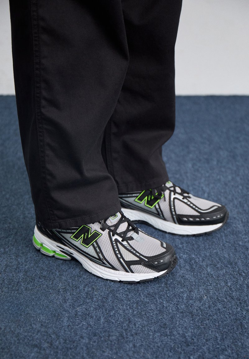Chaussures de sport grises et noires avec des accents verts, tige en maille et semelles rembourrées, associées à un pantalon noir sur un tapis bleu.