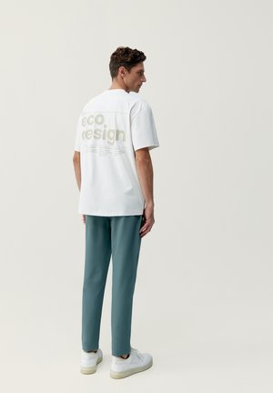 Bílý tričko s textem "eco.design" ve světle béžové na zádech, kombinované s tyrkysovými kalhotami a bílými teniskami. Jednoduchý, moderní design.