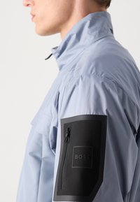 Light blue jacket with a high collar. Features a black zippered pocket on the left arm with a rectangular logo. Made of smooth, lightweight material.