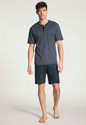 Camicia a maniche corte a righe blu navy e azzurre con taschino, abbinata a pantaloni corti blu navy, realizzati in tessuto morbido. Modello in piedi a piedi nudi.