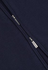 Granatowa bluza z zamkiem błyskawicznym, wykonana z teksturowanej tkaniny; posiada metalowy zamek i oznaczoną zawieszkę przy centralnym szwie.