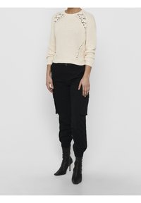 Karamellfärgad stickad tröja med spetsdetaljer vid axlarna, kombinerad med svarta cargobyxor. Modellen bär svarta klackskor, med händerna vid sidorna.