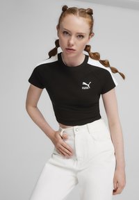 Must värviline lühikeste varrukatega ja lühikese lõikega T-särk, valged õlavarre aktsendid ja Puma logo ees, koos kõrge vöökohtadeta valgete pükste paariga.