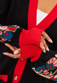 Černý cardigan s červeným lemem, zdobený květinovými pletenými vzory na rukávech. Hladké manžety, zapínání na knoflíky, měkká textura.