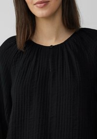 Czarna, fakturowana bluzka z fałdami, okrągłym dekoltem i małym guzikiem z przodu. Lekki materiał o lekko pomarszczonym wyglądzie.