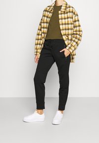 Camicia in flanella a scacchi gialla e nera, t-shirt verde a girocollo, pantaloni jogger neri e sneaker bianche. Completo casual con texture diverse.