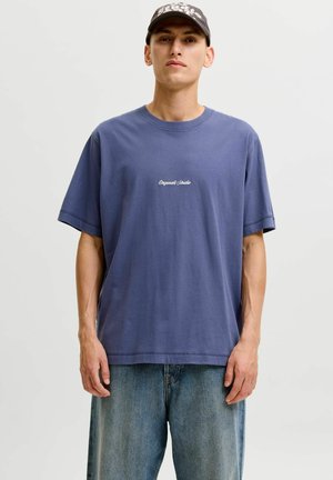 Jonge man draagt een blauwe oversized T-shirt met kleine witte tekst, losse spijkerbroek en een donkere pet, staand tegen een effen lichte achtergrond.