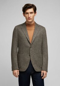 Brauner, strukturierter Blazer mit zwei vorderen Taschen und einem einzigen Knopfverschluss, über einem braunen Rollkragenpullover getragen.