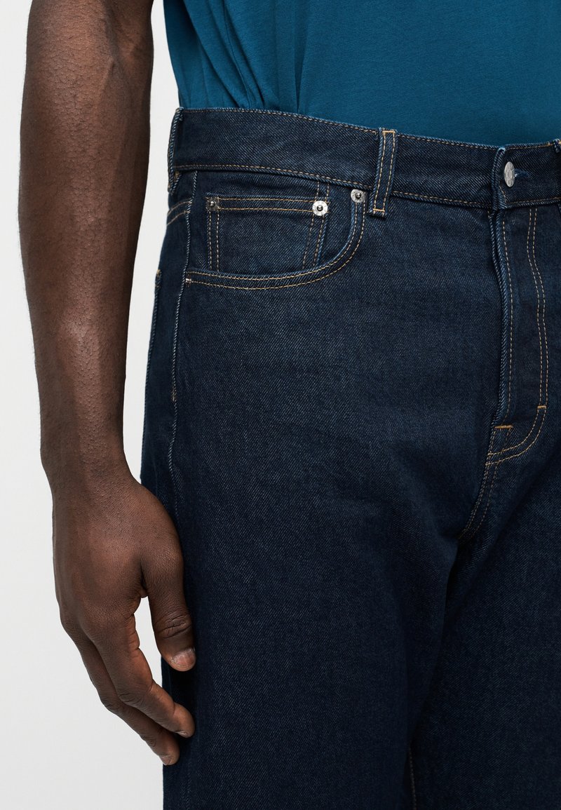 Dark-skinned person wearing dark blue denim jeans and a teal shirt, with their left arm relaxed by their side against a plain background.