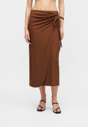 Brun midi nederdel i omslagsstil med snørredetalje i siden, båret med sorte åbne hæle-sandaler på en stående person.