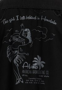 Zwarte stoffen shirt met een zilveren afbeelding van een hula danseres, tekst en een palmboom. Informatieve bedrukking bevat stationdetails en openingstijden.