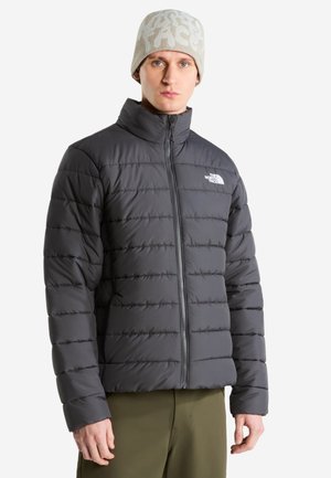 Hombre con chaqueta de plumón negra y gorro beige con dibujos, de pie con los brazos relajados frente a un fondo blanco.