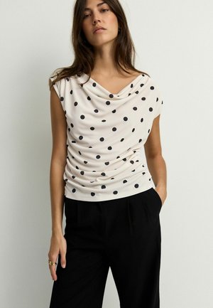 Halenka s názvem cowl neck, krátké rukávy, v krémové barvě s černými puntíky; měkká látka s řasením; spárováno s černými kalhotami.