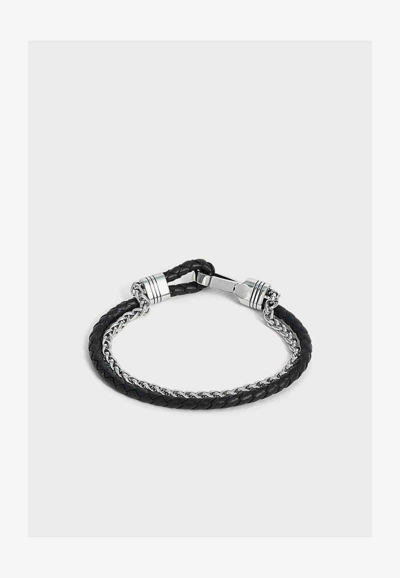 Bracelet en cuir tressé noir et chaîne en argent avec fermoir argenté sur un fond gris clair uni.
