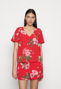 Top floral vermelho com decote em V e mangas curtas, apresentando flores cor-de-rosa e folhas verdes em um tecido suave. Combinado com uma saia vermelha correspondente.
