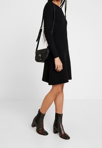 Schwarzes langärmliges Kleid mit einem ausgestellten Saum, kombiniert mit Knöchelstiefeln mit strukturiertem Reptilienmuster und Blockabsätzen sowie einer kleinen schwarzen Umhängetasche.