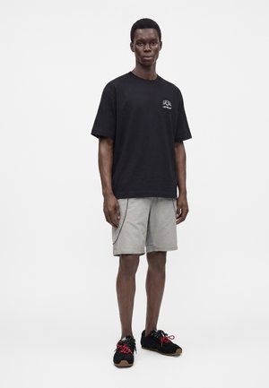 Νέος άνδρας στέκεται φορώντας μαύρο oversized μπλουζάκι, ανοιχτό γκρι σόρτς με μαύρο περιθώριο και μαύρα αθλητικά παπούτσια με κόκκινα κορδόνια σε λευκό φόντο.