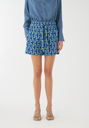 Femme portant un short bleu à motif taille haute avec ceinture, associé à un haut à manches longues assorti et des sandales vertes à lanières.