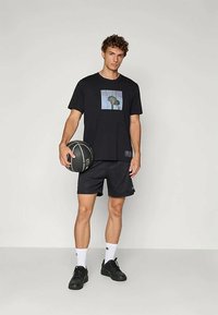 Czarna koszulka z graficznym nadrukiem, zestawiona z czarnymi krótkimi spodniami. Model trzyma piłkę koszykową, ubrany w czarne buty i białe skarpetki.