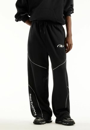 Personne portant un pantalon de survêtement noir New Balance avec logo blanc et passepoil blanc incurvé, assorti à des baskets noires et un hoodie noir.