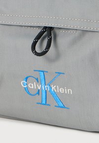 Γκρι υφασμάτινη θήκη με μαύρο λουρί, που φέρει το μπλε και λευκό κεντημένο λογότυπο "Calvin Klein" και τα αρχικά "CK".